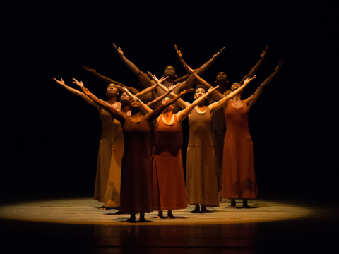 Alvin Ailey dancers on a dark stage raising their arms in the air and being lit by a single orange light.